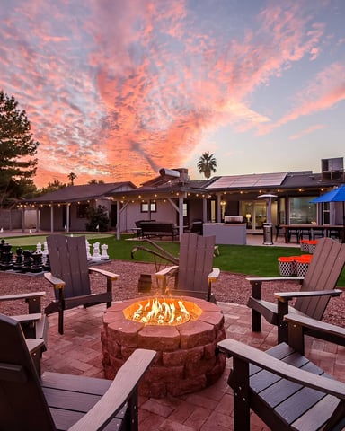 Backyard fire pit with Adirondack chairs, lit fireplace, lawn, and single-story home at sunset with dramatic pink sky