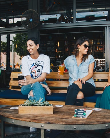 Four women sitting on wooden benches at an outdoor patio bar, holding drinks and conversing in front of large windows.