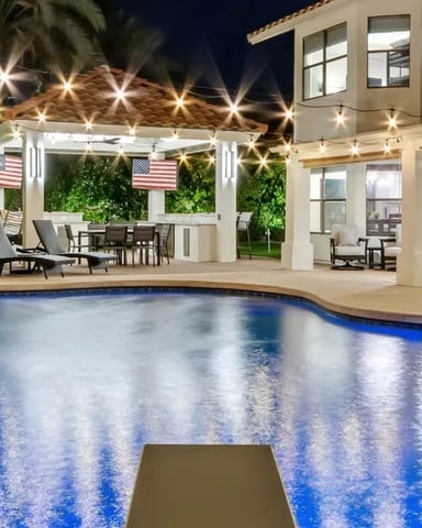 Illuminated backyard pool at night with beige house, pergola, lounge chairs, and string lights overhead