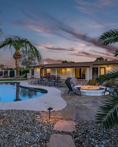 Backyard with curved pool, fire pit, lounge chairs, palm trees, and mid-century modern house at dusk