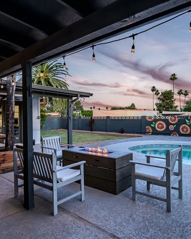 Covered patio with dining table overlooking pool, fire pit, and palm trees at sunset