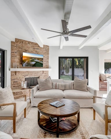 Bright open-plan living room with white ceiling beams, brick fireplace with TV, cream sofa, wooden chairs, round coffee table, and glass doors opening to patio.