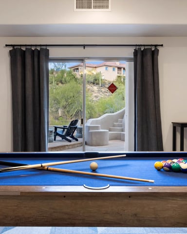 Game room with blue pool table, cues, and balls, glass doors opening to desert landscape and patio seating