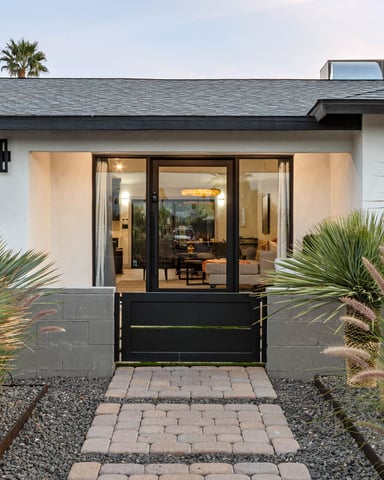 Modern white house exterior with black metal gate, glass doors, paver pathway, and desert landscaping with yucca plants
