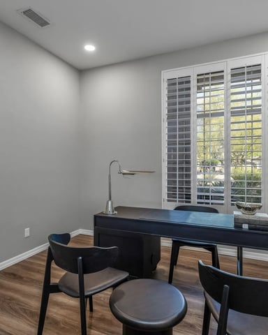 Home office with black desk, two black chairs, round ottoman, and white shuttered windows overlooking trees