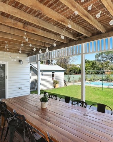 Covered timber deck with dining table overlooking backyard, pool, and white house with green lawn