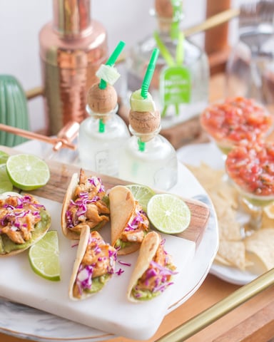 Styled table display with tacos topped with purple cabbage, lime wedges, cocktails with green straws, and decorative cacti