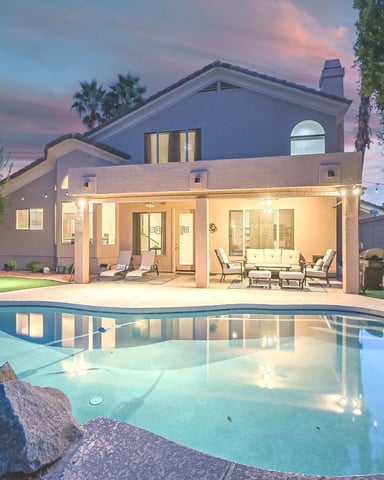 Modern house with lit patio and swimming pool at dusk, palm trees and manicured lawn visible in backyard