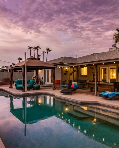 Backyard with illuminated mid-century home, pool, lounge chairs, covered patio, and palm trees at dusk