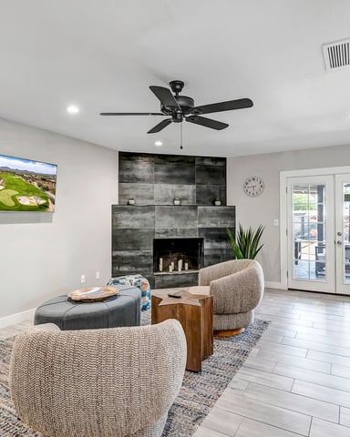 Open-plan living room with fireplace, ceiling fan, TV, upholstered chairs, and kitchen area with white cabinetry