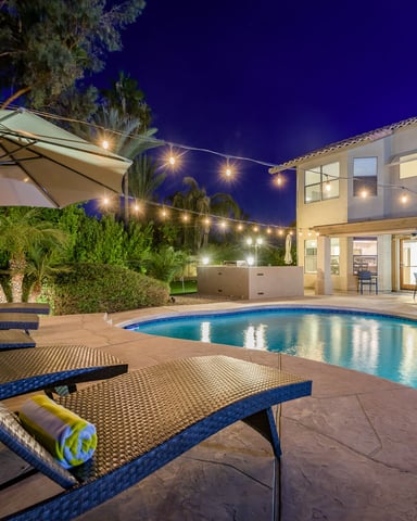 Illuminated backyard at night with swimming pool, lounge chairs, umbrella, and modern house with string lights