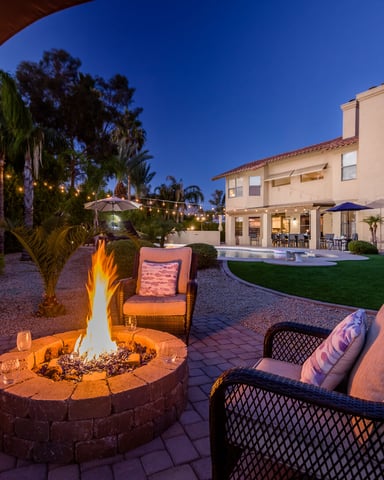 Backyard fire pit with lounge seating, pool, and two-story home at dusk with string lights