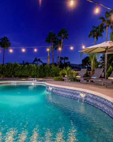 Illuminated resort pool at dusk with lounge chairs, umbrella, palm trees, and string lights overhead