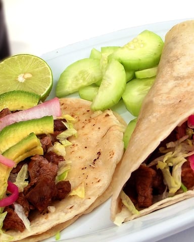 Two beef tacos with avocado, pickled onions, and cabbage on white plate with cucumber slices and lime wedge