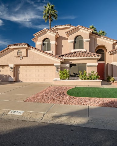 Southwestern-style two-story stucco house with tile roof, arched windows, and palm trees, desert landscaping with driveway.