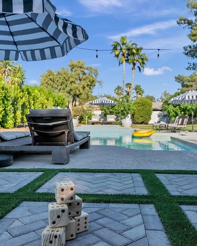 Backyard pool area with striped umbrellas, black lounge chairs, patio games, and palm trees under blue sky