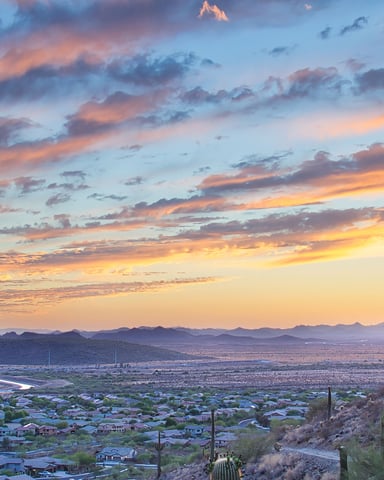 Sunset over desert valley with saguaro cacti, suburban development, and distant mountains under colorful sky