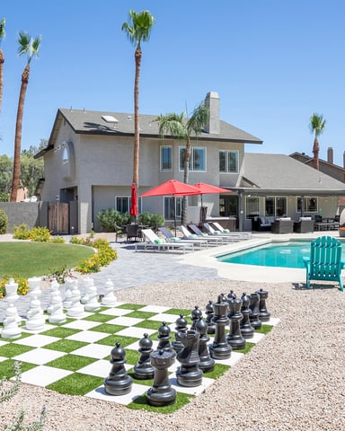 Desert home with pool, patio, red umbrella, lounge chairs, and oversized chess set in yard surrounded by palm trees