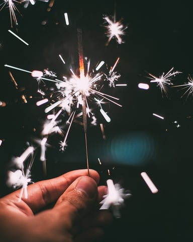 Hand holding a lit sparkler against dark night sky with sparks bursting outward