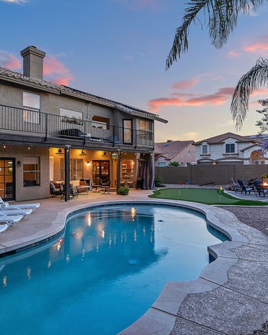 Backyard with illuminated pool, lounge chairs, two-story house with patio, putting green, fire pit, and palm trees at dusk