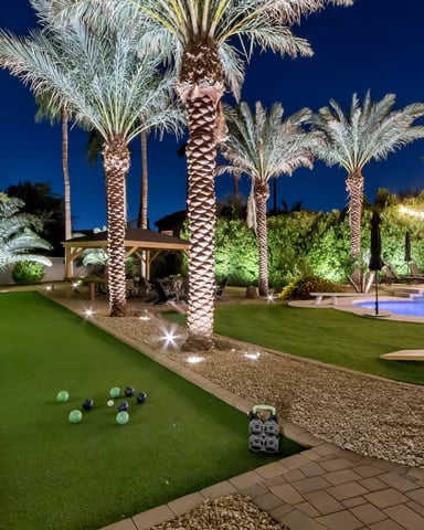 Nighttime backyard with illuminated palm trees, bocce ball court, swimming pool, lounge chairs, and landscaped gardens