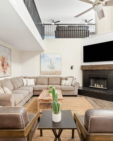 Modern two-story living room with beige sectional sofa, fireplace with TV above, wood furniture, and open staircase to upper level