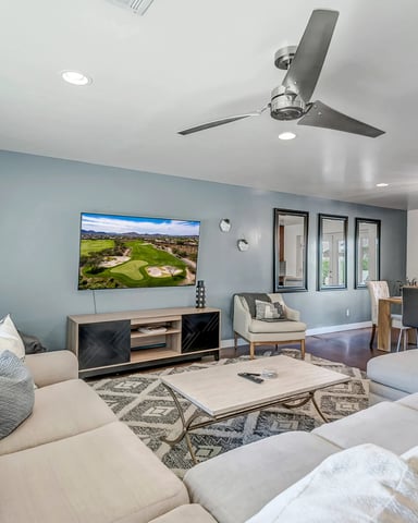 Open-plan living room with cream sectional sofa, blue accent wall, wall-mounted TV, and dining area with glass doors to outdoor patio.