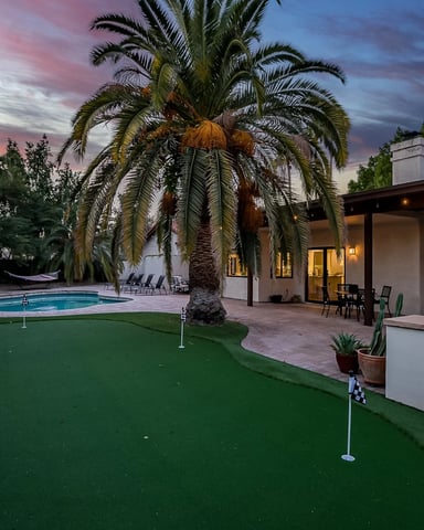 Backyard with putting green, pool, palm trees, and modern house at dusk with pink sky