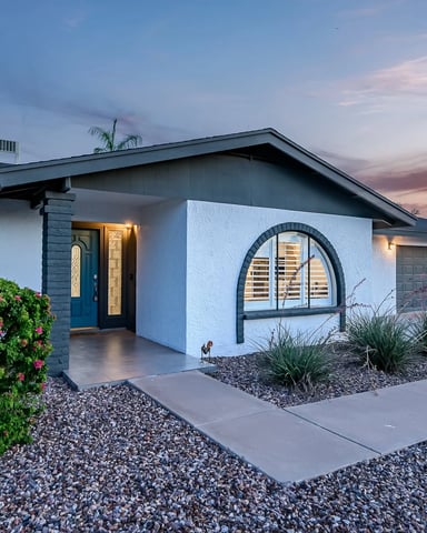 Modern single-story home with white and dark gray exterior, arched windows, blue front door, and desert landscaping at dusk