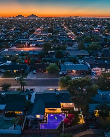 Aerial view of suburban neighborhood at dusk with illuminated houses, lit swimming pool in foreground, and mountains on horizon.