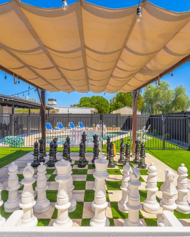 Backyard with large outdoor chess set, tan shade sail, pool with blue loungers, and black fencing
