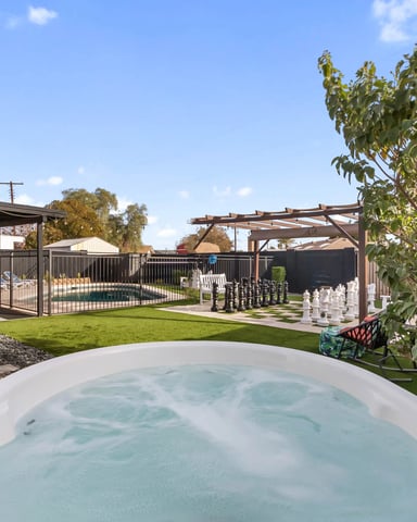 Backyard with hot tub in foreground, fenced pool, lawn, pergolas, and covered patio area under clear sky