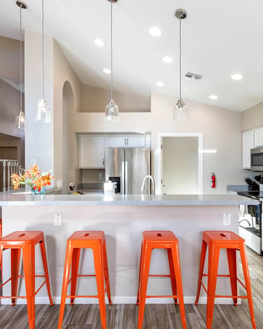 Modern kitchen with white cabinetry, stainless steel appliances, and island with four orange metal bar stools, adjacent to staircase