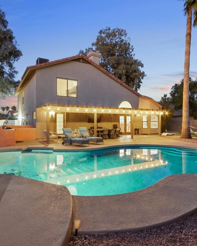 Backyard with illuminated swimming pool, lounge chairs, pergola with curtains, and house at dusk surrounded by palm trees
