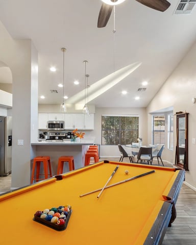 Open-plan recreation room with yellow pool table, kitchen with white cabinets, orange bar stools, and glass doors opening to backyard