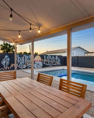 Covered patio with wooden dining table and chairs, string lights overhead, pool and desert mural wall visible at dusk