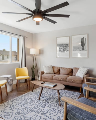 Bright living room with tan sofa, yellow chairs, ceiling fan, and desert landscape artwork on white walls