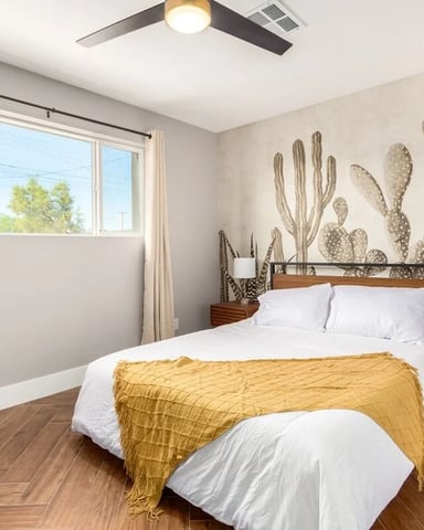 Bedroom with wood bed frame, white bedding, yellow throw blanket, large cactus wall mural, wood nightstand, and ceiling fan.