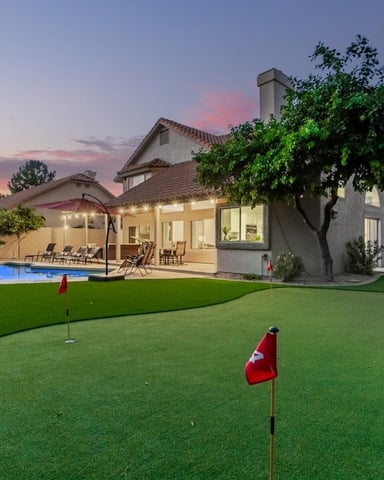 Backyard with putting green, illuminated pool, patio seating, and two-story house at dusk