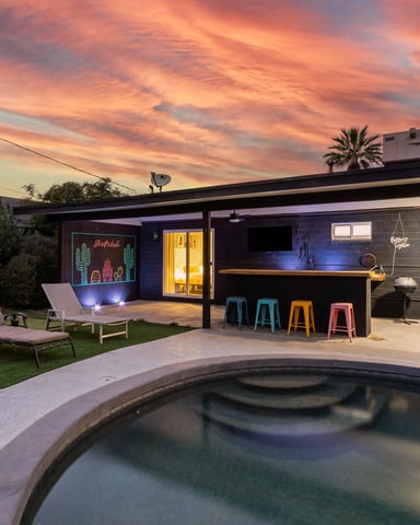 Backyard with pool, lounge chairs, outdoor bar with colorful stools, and modern house at sunset
