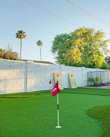 Backyard mini golf course with artificial turf, red flags, and cornhole boards against white block wall