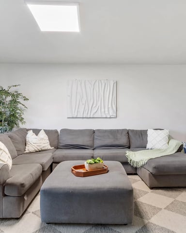 Modern living room with large gray sectional sofa, ottoman coffee table, potted plant, and floor mirror overlooking kitchen