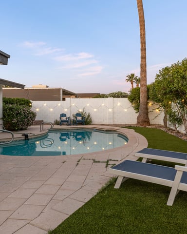 Backyard with round swimming pool, lounge chairs, palm tree, white fence, and modern house with patio