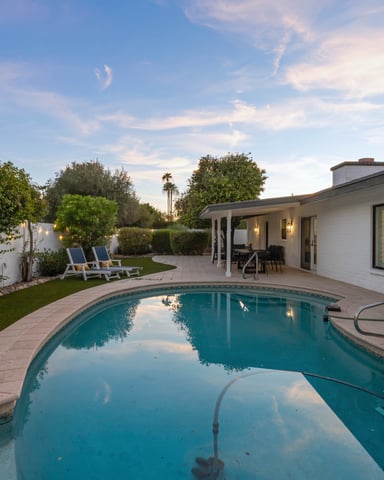 Mid-century modern house with curved swimming pool, patio seating, manicured lawn, and desert landscaping at dusk