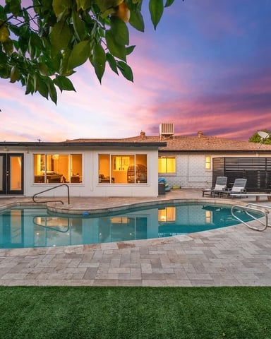 Backyard with illuminated mid-century home, swimming pool, patio seating, and sunset sky