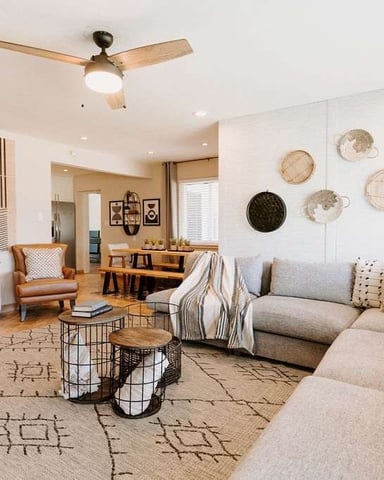 Open-plan living room with tan sectional sofa, cognac leather chairs, woven basket wall decor, and ceiling fan.
