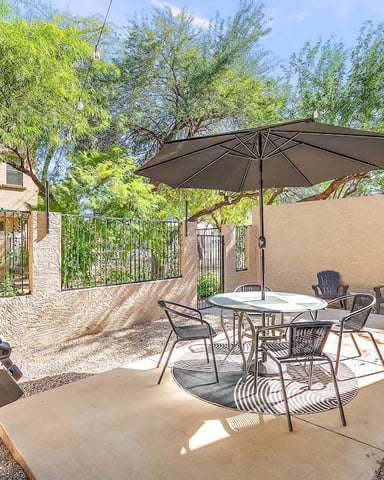 Desert backyard patio with dining table, umbrella, lounge chairs, grill, and pool under shade trees