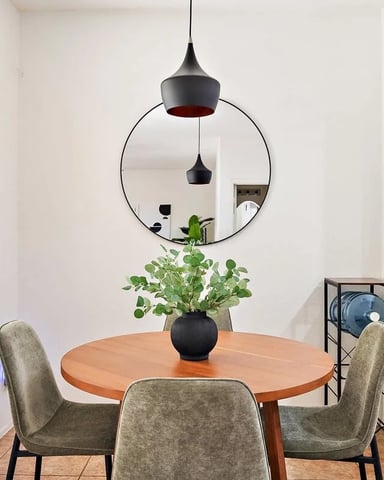 Dining room with round wood table, three upholstered chairs, black pendant lights, circular mirror, and potted plant centerpiece.