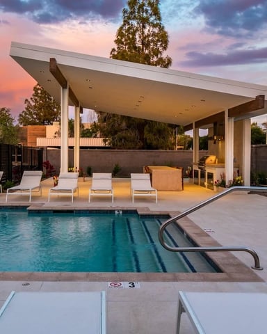 Modern backyard pool with lounge chairs under covered patio at sunset, palm trees visible in background