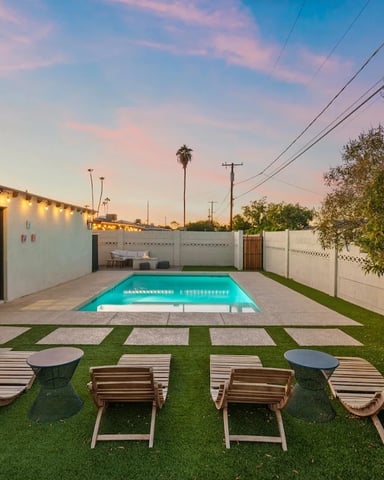 Backyard with rectangular swimming pool, lounge chairs, white fence, and illuminated house at dusk
